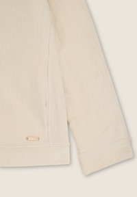 Manica di felpa in corduroy beige con texture a costine, dotata di un piccolo etichetta metallica vicino all'orlo e una finitura liscia.
