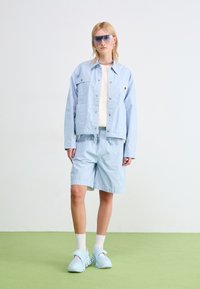 Giacca di jeans a righe azzurre con bottoni a scatto e tasche, abbinata a pantaloni corti coordinati. Gli accessori includono occhiali da sole e scarpe azzurre.