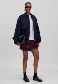 Marineblauwe oversized bomberjack, wit T-shirt, rode geruite minirok. Zwart leren loafers en sokken. Een clutch en zonnebril maken de look compleet.