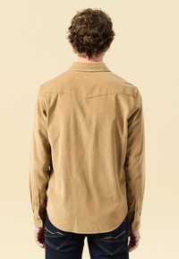Camicia di velluto a coste color tan con colletto a punta, maniche lunghe e polsini con bottone; presenta un orlo arrotondato e un design con yoke posteriore.