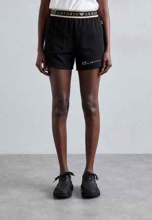 Person trägt schwarze Emporio Armani Shorts und schwarze Sneakers und steht auf einem grauen Boden vor einer grauen Wand.