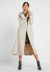 Beige trenchcoat med dubbelknäppning, långa ärmar och längd till mitten av vaden. Har knappar och ett slätt, texturerat tyg.