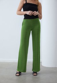 Kvinna klädd i högmidjade gröna vida byxor, ärmlös svart topp med rynkor, svarta klacksandaler och guldringar på ett betonggolv inomhus.