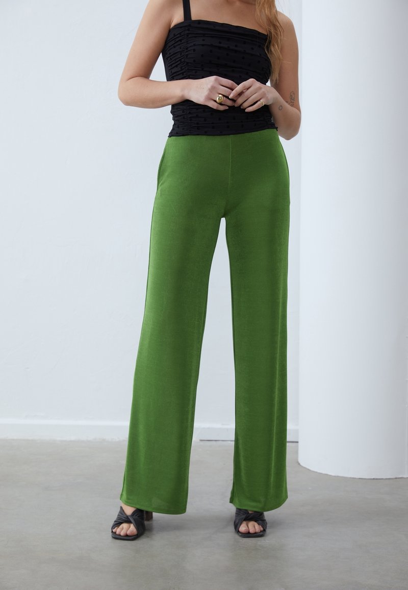 Kvinna klädd i högmidjade gröna vida byxor, ärmlös svart topp med rynkor, svarta klacksandaler och guldringar på ett betonggolv inomhus.