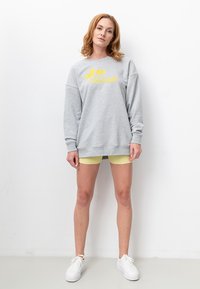 Hellgrauer Pullover mit gelbem Drucktext, kombiniert mit gelben Rippenshorts und weißen Sneakers. Lässiger, lockerer Schnitt.