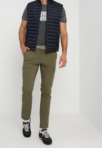 Marineblå puffervest over en grå t-shirt, olivengrøn cargo-bukser og sorte sneakers. Vesten har en høj krave og vandret syning.