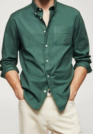Homme portant une chemise boutonnée vert foncé à manches longues retroussées et un pantalon couleur crème, mains dans les poches.