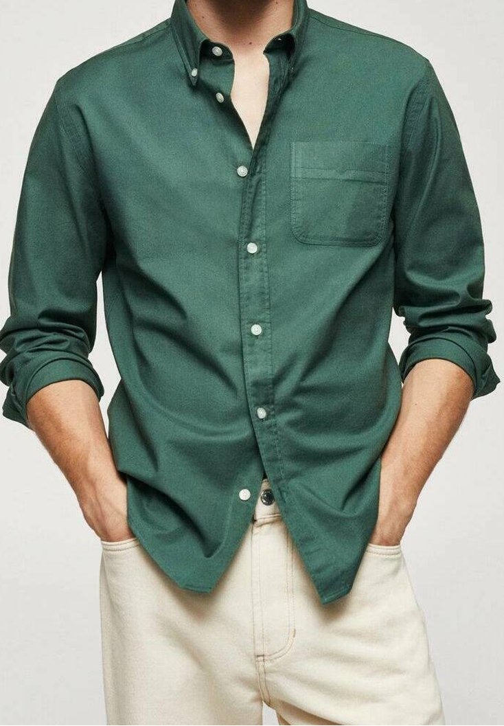 Homme portant une chemise boutonnée vert foncé à manches longues retroussées et un pantalon couleur crème, mains dans les poches.