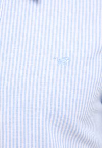 Camicia a righe blu chiaro e bianca in cotone, con un logo di un animale ricamato sul petto. Il tessuto ha una consistenza liscia.