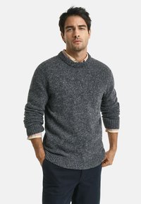 Mann steht mit den Händen in den Taschen und trägt einen gesprenkelten dunkelgrauen Strickpullover über einem hellen karierten Hemd und dunklen Hosen.