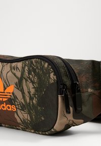 Boková taška s kamuflážním vzorem, vyrobená z odolné látky, se dvěma zipovými přihrádkami a oranžovým logem Adidas na přední straně.