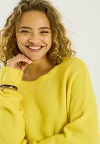 Mujer joven con cabello rizado sonriendo, vestida con un suéter amarillo brillante y pulseras, posando con la mano cerca del mentón contra un fondo simple.