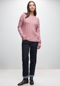 Lichtroze kabelgebreide trui met een textuurpatroon, geribbelde mouwen, gedragen over donkere jeans met opgerolde pijpen enbruine enkellaarzen.