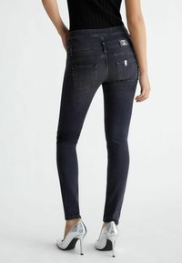 Jeans de mezclilla negra ajustada con una cinturilla de altura media, diseño de cinco bolsillos y detalles de costura sutiles. Llevados con zapatos de tacón alto plateados.