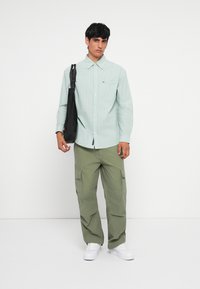 Hellgrünes gestreiftes Hemd mit Knöpfen und Brusttasche, kombiniert mit olivgrünen Cargo-Hosen und weißen Turnschuhen. Schwarze Umhängetasche auf der linken Seite.
