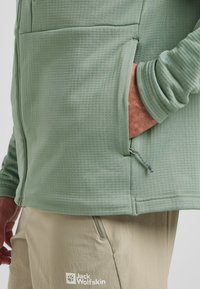 Veste en polaire vert clair avec un motif texturé, dotée d'une poche latérale et d'un cordon de serrage ajustable. Pantalon beige avec logo visible en bas.