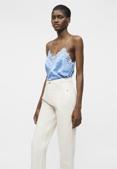 Ženska, ki nosi svetlo modro satenasto majico s čipkastim robom in tankimi naramnicami ter visokim pasom belo-bele hlače na enobarvnem ozadju.