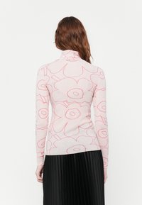 Prilagođeni turtleneck pulover u svijetlo ružičastoj boji s odvažnim crvenim cvjetnim uzorkom, dugih rukava i glatke teksture. Ispod se nalazi plisirana crna suknja.