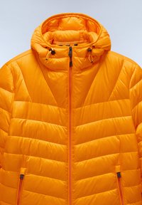 Giacca arancione imbottita con collo alto e cappuccio regolabile, caratterizzata da cuciture diagonali e una cerniera frontale con una piccola tasca laterale.