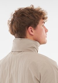 Giacca a vento beige con colletto alto e tessuto testurizzato, vista di lato, che mostra il colletto e i capelli della persona che la indossa.