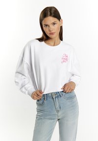 Witte sweatshirt met lange mouwen, verlaagde schouders en roze geborduurde tekst op de borst. Draagt een lichtblauwe high-waisted jeans.