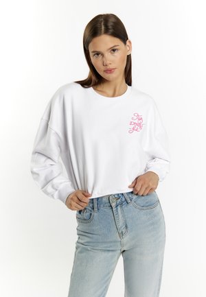 Witte sweatshirt met lange mouwen, verlaagde schouders en roze geborduurde tekst op de borst. Draagt een lichtblauwe high-waisted jeans.