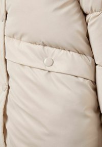 Beige pufferjakke-sektionen med polstret tekstur og glat overflade. Har en fremtrædende vandret søm og en rund trykknapsdetalje.