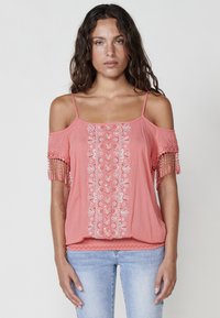Koralo spalvos off-shoulder palaidinė su išsiuvinėjimais gėlių raštais ir frakcinėmis rankovėmis. Pagaminta iš lengvos medžiagos, pasižymi laisvu siluetu ir banginiais kraštais.