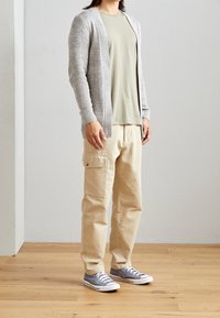 Hellgrauer gestrickter Cardigan über einem hellgrünen Hemd, kombiniert mit beigen Cargo-Hosen und grauen Sneakers mit weißen Sohlen. Einfaches Design und Passform.