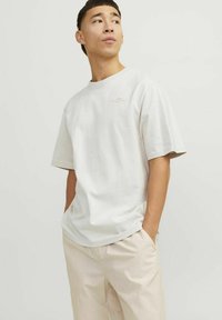Light gray short-sleeve t-shirt, round neckline, soft cotton fabric, subtle logo detail on chest, paired with beige trousers.