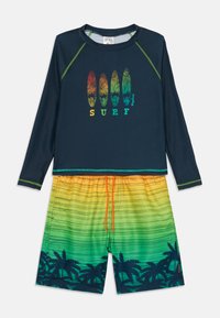 Dlhý rukáv námorníckej modrej rash guard s zeleným prešívaním, s grafikou surfboardu. Šortky so žlto-zeleným gradientom a vzorom palmových stromov s šnúrkou.
