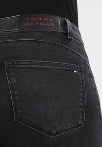 Jeans de mezclilla negra que presentan un parche de cuero en la cintura con "TOMMY HILFIGER" grabado en rojo. El bolsillo trasero tiene un pequeño detalle del logotipo tricolor.