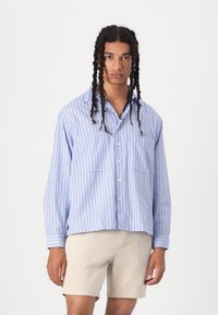 Camicia blu a maniche corte con bottoni, strisce bianche verticali, due tasche sul petto e una vestibilità comoda. Indossata con pantaloni corti beige chiaro.