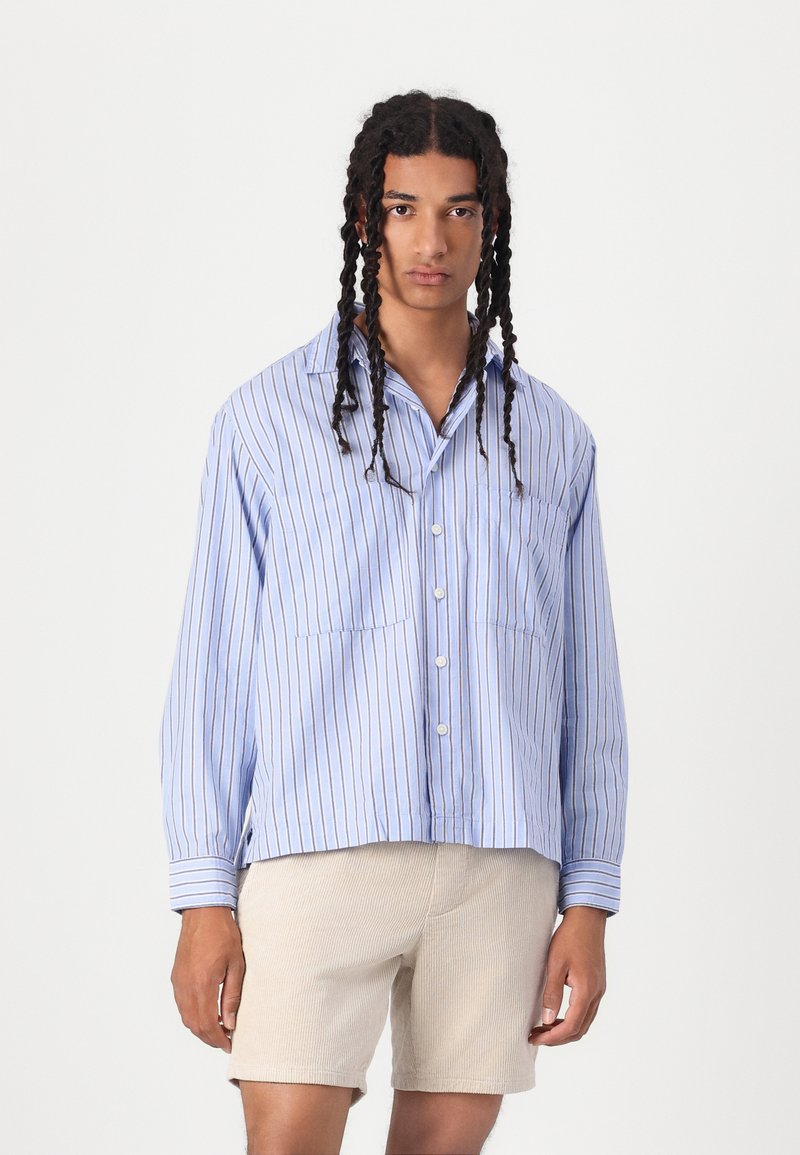 Camicia blu a maniche corte con bottoni, strisce bianche verticali, due tasche sul petto e una vestibilità comoda. Indossata con pantaloni corti beige chiaro.