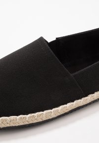 Gros plan d'une chaussure noire en tissu sans lacets avec une bordure en jute beige tressée le long du bord de la semelle.
