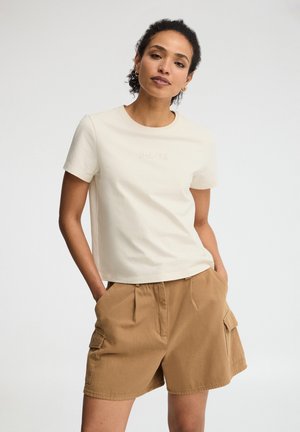 Donna con capelli ricci che indossa una maglietta corta color crema e pantaloncini cargo beige, in piedi con le mani in tasca davanti a uno sfondo semplice.
