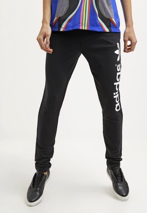 Osoba nosząca czarne spodnie dresowe Adidas z białym logo na nodze i czarne trampki, stojąca na jednolitym tle.