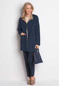 Marineblauer Woll-Mix-Mantel mit großen Fronttaschen, beigem Kragen und Knopfverschluss. Kombiniert mit marineblauen Hosen und braunen Slippern.