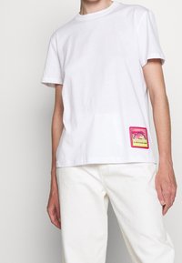 Camiseta de algodón blanca con mangas cortas, escote redondeado y un parche rectangular de logo en rosa y amarillo en el lado izquierdo.