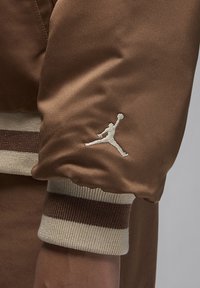 Brązowa satynowa rękaw kurtki z białym haftowanym logo Jumpman oraz ściągaczem z kontrastującymi beżowymi i ciemnobrązowymi paskami.