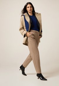 Beige trenchcoat over een blauwe trui, beige op maat gemaakte broek en zwarte enkellaarsjes met hak; heeft op maat gemaakte snits en een elegante silhouet.