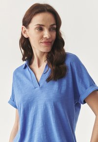 Polo shirt blu a maniche corte con scollo a V, texture morbida e maniche arrotolate. Realizzato in materiale leggero con una leggera lucentezza.