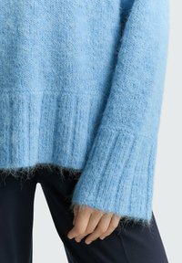 Jasnoniebieski, puszysty sweter z prążkowanym dołem i mankietami; miękka tekstura; luźny fason; ukazujący rękę spoczywającą na ciemnych spodniach.