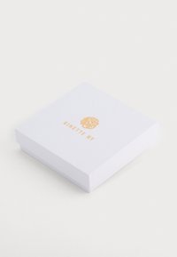 Baltas kvadratinis dovanų dėžutė su tekstūruotu paviršiumi, ant viršaus esantis auksinių raidžių iškilmingas "GINETTE NY" logotipas.