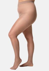 Persona incinta che indossa collant premaman trasparenti color carne, in piedi con un piede sollevato su uno sfondo neutro.
