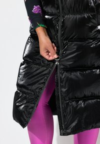 Schwarze Puffer-Weste mit glänzender Textur und Frontreißverschluss, getragen über lila Leggings. Hand, die den Reißverschlussgriff zum Schließen greift.
