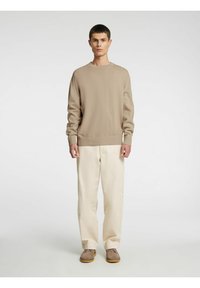 Beige strikkegenser med rund hals, kombinert med kremfargede bukser. Modellen har på seg lysebrune sko. Enkelt og minimalistisk design.