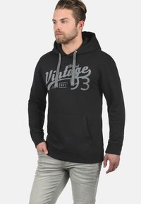 Sudadera con capucha negra, con un bolsillo frontal y un estampado gráfico vintage en gris. Tejido suave, corte holgado y puños y dobladillo acanalados.