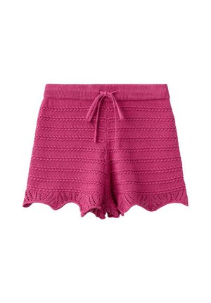 Kvinde med langt sort hår iført hvid stropløs top, lyserøde strikkede shorts, blå klipklapper, stående med den ene hånd på hoften mod en ensfarvet væg.
