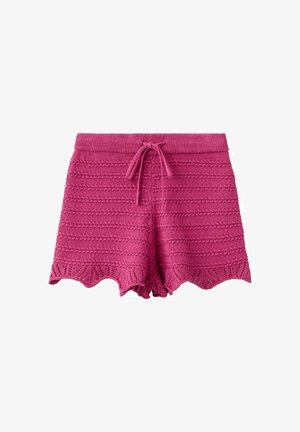 Kvinde med langt sort hår iført hvid stropløs top, lyserøde strikkede shorts, blå klipklapper, stående med den ene hånd på hoften mod en ensfarvet væg.